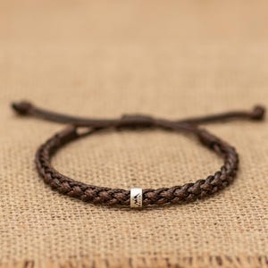 May include: A dark brown braided cord bracelet featuring a single cylindrical silver-toned metal bead. The bead is engraved with a small, simple black mountain range graphic. The bracelet has adjustable cord ends extending from the main band.