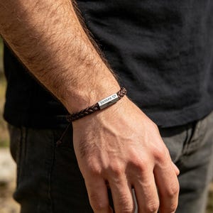 Op de afbeelding: Gevlochten bruine armband met een zilveren staafje gegraveerd met de naam "James" en een hartsymbool. De armband is gemaakt van geweven materiaal en heeft een verstelbare sluiting. Een stijlvol accessoire.
