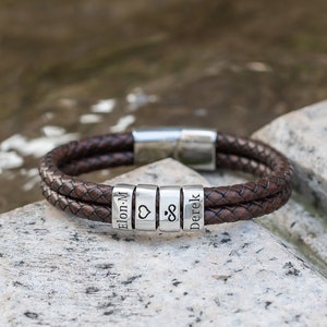Pode incluir: Um bracelete de couro castanho com contas de prata. As contas são gravadas com os nomes "Elon.M", um símbolo de coração, um símbolo de infinito e "Derek".