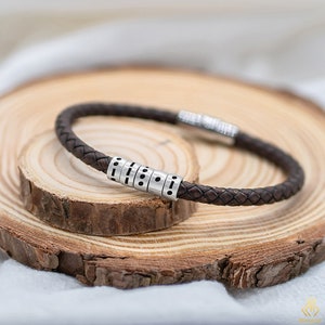 Puede incluir: Un brazalete de cuero marrón con cuentas de plata. Las cuentas tienen un patrón de puntos y líneas.