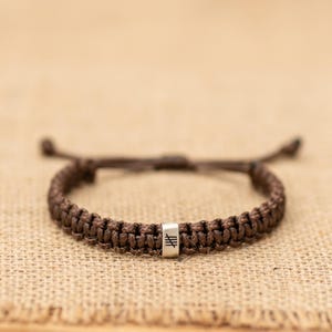 May include: A brown woven cord bracelet featuring a central silver-toned metal bead. The bead is engraved with a black symbol consisting of three horizontal lines crossed by a single vertical line. The bracelet has adjustable cord ends.