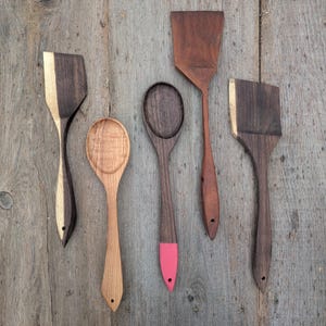 May include: A collection of five wooden kitchen utensils, including a spatula, spoon, and turner. The utensils are made of various types of wood, with some featuring contrasting colors and painted accents. The background is a rustic wooden surface.