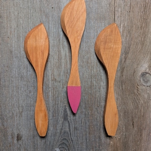 May include: Three wooden spoons with a leaf-shaped head and a slender handle. One spoon has a pink painted tip. The spoons are made of light-colored wood and are arranged on a gray wooden surface.