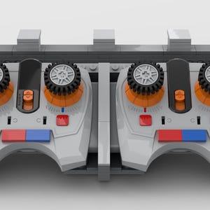 May include: A grey and white toy controller with orange and black detailing. The controller features two joysticks with gear-like wheels and red and blue buttons. The symmetrical design suggests it is for a vehicle or machine.