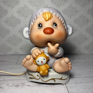 May include: A ceramic lamp shaped like a baby wearing a white bonnet and outfit, holding a small yellow monkey toy. The baby has a round orange nose, rosy cheeks, and blonde hair. The base is white with gold filigree accents and a power cord.