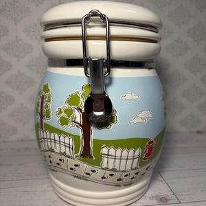 May include: A white ceramic canister with a metal clamp lid featuring a colorful illustration of a suburban scene with green trees, a white picket fence, a red fire hydrant, and a sidewalk with black paw prints against a light blue sky.