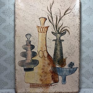 May include: A rectangular ceramic wall plaque with a speckled cream background featuring a painted still life of three vases and a bowl. The artwork displays a tall yellow bottle, a dark grey vase with sprigs, and a blue bowl, signed Freeman.