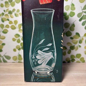 May include: A rectangular cardboard box featuring a clear glass vase with a flared rim and a white etched floral design. The box is dark green with a red plaid ribbon graphic at the top and includes the text Vase Décoré de 18 cm at the base.