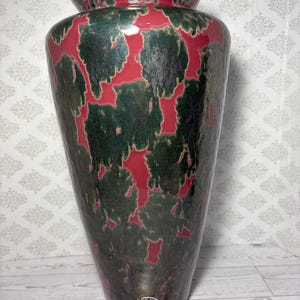 May include: A tall, tapered ceramic vase featuring a glossy finish with a mottled pattern of deep forest green and dark charcoal over a vibrant red base. A small circular sticker with illegible text is affixed near the bottom of the vase.