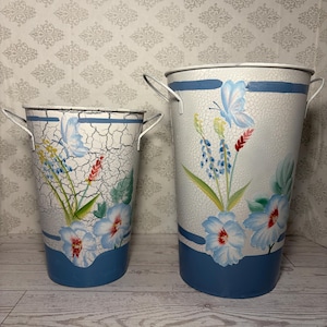May include: Two tapered metal buckets with side handles, featuring a white crackle finish, blue horizontal bands, and hand-painted floral designs including blue flowers, green leaves, and a butterfly on each container in varying sizes.