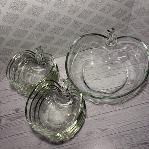 May include: A set of clear glass serving dishes shaped like apples, including one large bowl and several smaller matching bowls. Each piece features a molded stem detail at the top, a smooth interior, and a curved, rounded silhouette made of glass.