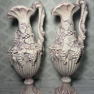 May include: A pair of ceramic pitchers featuring a raised relief design of grape clusters and vine leaves. The pitchers have a cream-colored base with purple-toned accents, a fluted lower body, a pedestal base, and a curved handle on the side.