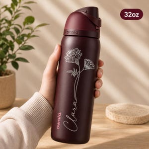 May include: A maroon insulated water bottle with a flip-top lid, featuring a floral design and the name "Clara" in cursive. The bottle has a capacity of approximately 946ml and the brand name "owala" is visible.