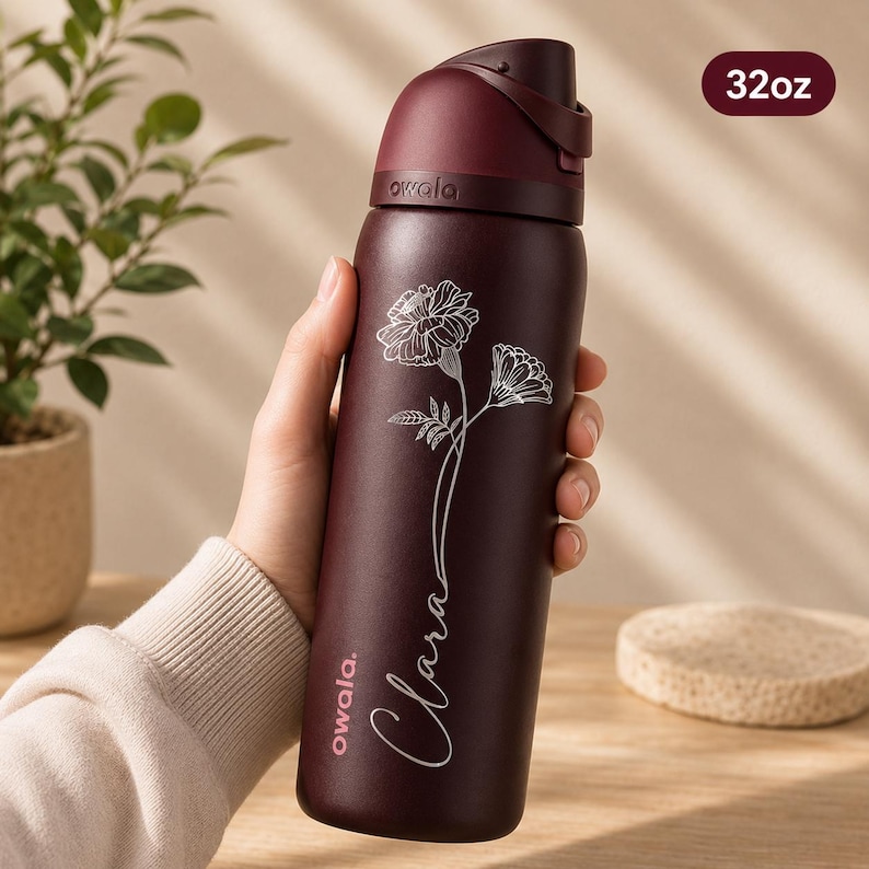May include: A maroon insulated water bottle with a flip-top lid, featuring a floral design and the name "Clara" in cursive. The bottle has a capacity of approximately 946ml and the brand name "owala" is visible.