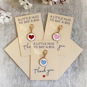 May include: Three gold-toned metal charms with lobster clasps, each featuring a circular enamel heart in red, pink, or light blue. Each charm is attached to a cream-colored card printed with the text A LITTLE HUG TO SAY A BIG Thank you.