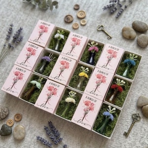 May include: A rectangular box containing ten individual compartments, each holding a small glass bottle with a miniature floral arrangement. Each compartment features a pink sleeve printed with the text A BUNCH OF love and a floral illustration.
