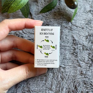 May include: A small white card with the text "BENEFITS OF BOX BREATHING" and an illustration of a turtle. The card explains the box breathing technique, including inhale, hold, and exhale steps. The card is held by a person.