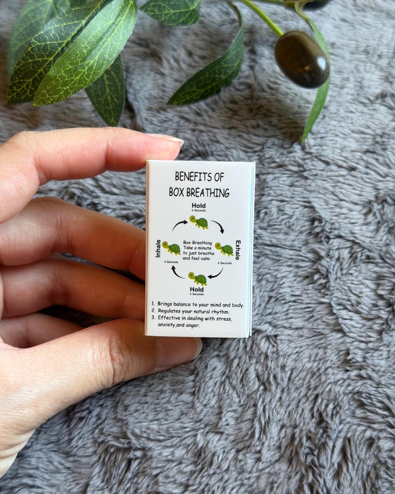 May include: A small white card with the text "BENEFITS OF BOX BREATHING" and an illustration of a turtle. The card explains the box breathing technique, including inhale, hold, and exhale steps. The card is held by a person.