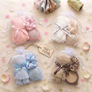 May include: Four sheer organza gift bags containing a plush textured washcloth, a matching satin scrunchie, and a round compact mirror. Each set is tied with a ribbon, and a wooden tag labeled GIFT FAVOR sits in the center among scattered petals.