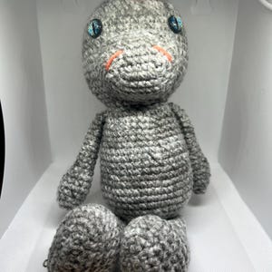 May include: A handmade gray crochet dinosaur plush toy with blue eyes and orange accents. The dinosaur has a small orange crest on its head and is sitting with its legs extended. The toy is made of soft yarn.