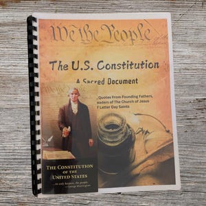 May include: A book titled "We the People: The U.S. Constitution, A Sacred Document" with a portrait of a man in a brown suit holding a quill pen. The book cover features a quote from Founding Fathers, leaders of The Church of Jesus Christ of Latter-day Saints.