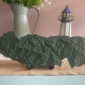 May include: A dark green, textured, topographical map sculpture, mounted on a black stand. A decorative lighthouse and a green jug with flowers are in the background. The map's surface has a detailed, crinkled appearance.