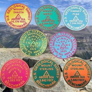 May include: A collection of eight circular U.S. Geological Survey benchmark markers in various colors including yellow, teal, light blue, green, pink, and orange, each featuring mountain names, elevations in feet, and a central triangle symbol.