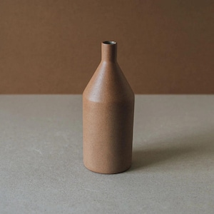May include: A cylindrical ceramic vase with a narrow neck and a matte, speckled light brown finish. The vessel features a flat base and a simple, uniform structure with a smooth texture, showcasing a consistent earthy tone throughout the entire body.