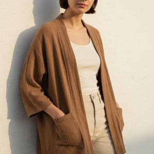 Op de afbeelding: Een bruin vest met open voorkant en twee voorzakken. Het vest wordt gedragen over een witte tanktop en gecombineerd met een beige broek. De stof heeft een getextureerde uitstraling. Het model staat tegen een witte muur.