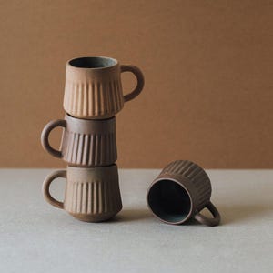 Puede incluir: Tres tazas de cerámica marrón apiladas, con detalles acanalados verticales y asas pequeñas. Una taza está tumbada de lado. Las tazas tienen un acabado mate y un interior oscuro.