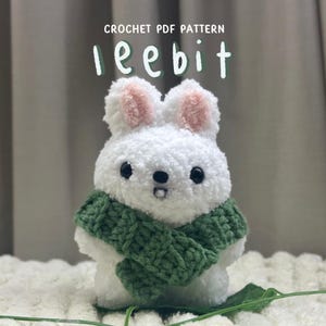 May include: A white crocheted bunny wearing a green crocheted scarf. The bunny has pink ears and black eyes. The text "CROCHET PDF PATTERN lee bit" is above the bunny.
