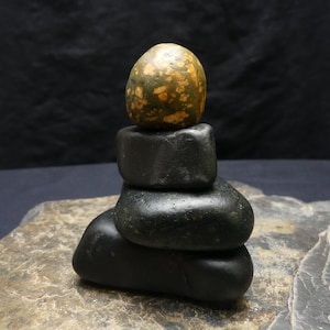 May include: A stack of four smooth, rounded stones arranged vertically. The top stone is mottled green and tan, while the three stones beneath it are solid dark charcoal grey. The entire stack rests upon a flat, textured, grey stone base.