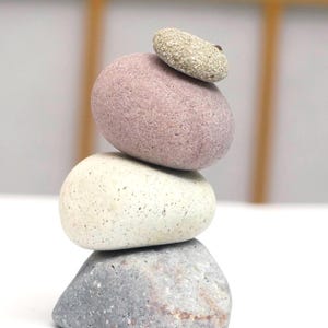 May include: A balanced stack of four smooth, rounded stones in shades of grey, white, and pink. The stones are stacked on top of each other, forming a small tower. The smallest stone is at the top, with the largest at the base.