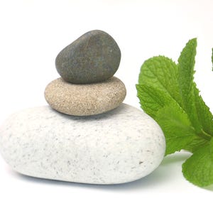 May include: A stack of three smooth, rounded stones arranged vertically. The bottom stone is large and white with grey speckles, the middle stone is tan and textured, and the top stone is dark grey. A sprig of fresh green mint leaves sits beside them.