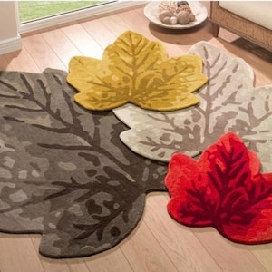 Op de afbeelding: Een verzameling bladvormige vloerkleden in diverse kleuren, waaronder bruin, geel, rood en beige. De kleden liggen op een houten vloer en tonen gedetailleerde bladnervenpatronen. Deze decoratieve kleden voegen een vleugje natuurlijke decoratie toe.