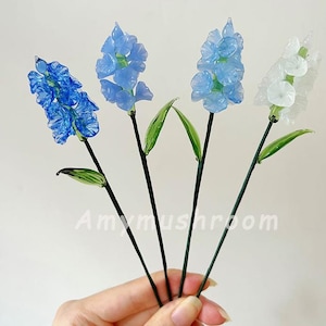 Puede incluir: Cuatro tallos de flores de vidrio con flores de vidrio azules y blancas. Las flores están dispuestas en un ramo y se sostienen en una mano.