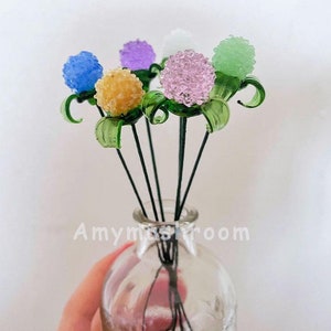 May include: A bouquet of six glass flowers in a clear glass vase. The flowers are in shades of blue, purple, pink, yellow, and green. The flowers are all round and have a textured surface.