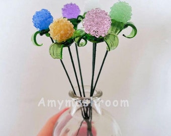 Puede incluir: Un ramo de seis flores de vidrio en un jarrón de vidrio transparente. Las flores son en tonos de azul, morado, rosa, amarillo y verde. Las flores son todas redondas y tienen una superficie texturizada.
