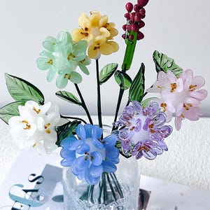 May include: A glass flower arrangement in a clear vase. The flowers are various colours, including white, blue, yellow, green, and pink. The arrangement includes green leaves and stems. The vase sits on a magazine with the text "IDEAS" and "Amymushroom".