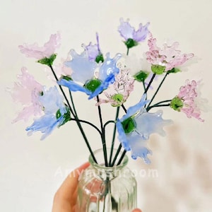 Może przedstawiać: Bukiet szklanych kwiatów w odcieniach niebieskiego, różowego i fioletowego. Kwiaty są ułożone w przezroczystym szklanym wazonie z fakturowaną powierzchnią.