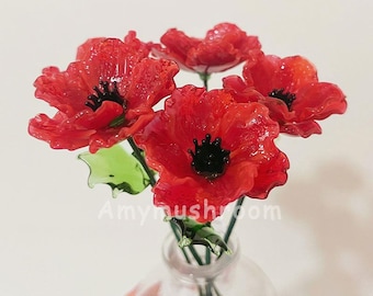 May include: A bouquet of red glass poppies with black centers in a clear glass vase. The poppies are arranged in a cluster and have a textured, realistic appearance.