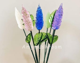 May include: A bouquet of five glass flowers in a clear glass vase. The flowers are in shades of white, pink, blue, and purple. The flowers have a textured, bubbly appearance. The stems are green and the leaves are green.