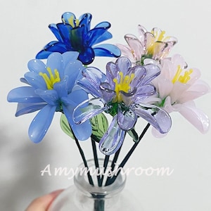 Puede incluir: Un ramo de flores de cristal en tonos azules, morados y rosas, con centros amarillos y hojas verdes. Las flores están dispuestas en un jarrón de cristal transparente. El texto "Amymushroom" es visible.