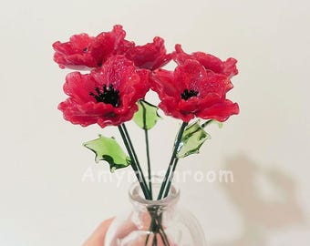 May include: A bouquet of five red glass poppies with green glass leaves in a clear glass vase.