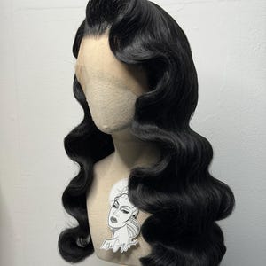 May include: A black, vintage-style wig with long, wavy curls. The wig is styled with a side part and features a natural-looking hairline. The wig is displayed on a mannequin head with a sticker that says "The Guy Hair".