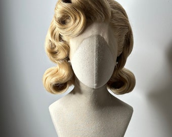 Może przedstawiać: Blond peruka w stylu vintage z lokami, prezentowana na głowie manekina. Peruka ma jasny, złoty kolor i jest stylizowana z dużymi, zdefiniowanymi lokami. Manekin ma neutralną maskę na twarz.
