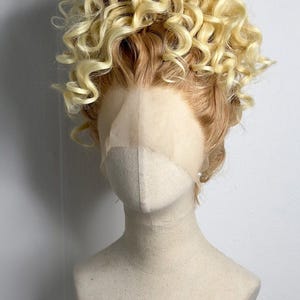 Könnte beinhalten: Blond und hellbraune Lockenperücke auf einem Schaufensterkopf. Die Perücke hat eine Spitzenfront und einen voluminösen, strukturierten Stil mit einer Mischung aus hellen und dunkleren Blond- und Brauntönen. Die Schaufensterpuppe ist neutral gefärbt.