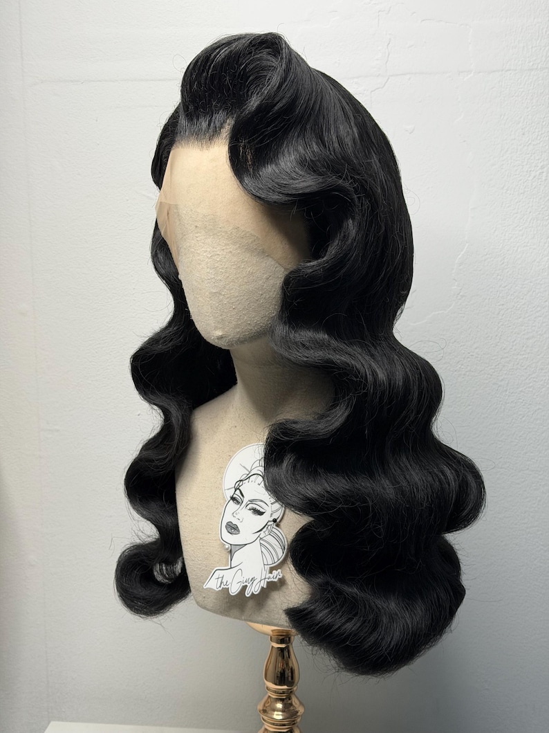 May include: A black, vintage-style wig with long, wavy curls. The wig is styled with a side part and a high, voluminous wave at the front. The wig is displayed on a mannequin head with a sticker that says "The Cozy Hair".