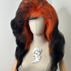 Könnte beinhalten: Ein Schaufensterpuppenkopf, der eine lange, wellige Perücke mit einem leuchtend orangefarbenen Oberteil und einem schwarzen Ombre zeigt. Eine weiße Anhänger-Halskette mit einem stilisierten weiblichen Gesicht hängt an der Schaufensterpuppe. Die Perücke hat einen Seitenscheitel und lange, fließende Schichten.