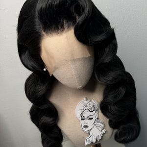 May include: A black wig with voluminous, wavy curls styled in a vintage Hollywood fashion. The wig is displayed on a mannequin head with a lace front. A small tag with a graphic of a woman's face is attached to the mannequin.